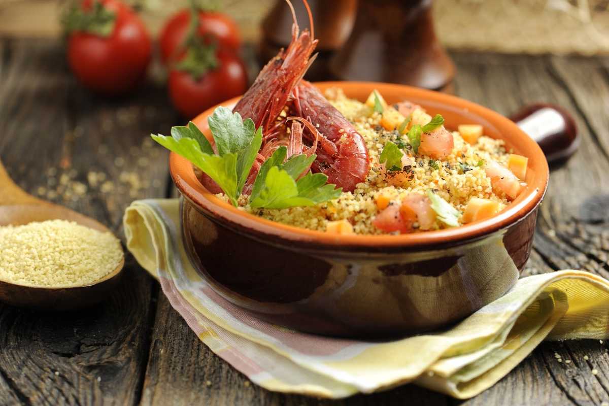Scorcio di una tavolata all’aperto con piatti tipici siciliani (cous cous di pesce, busiate, panelle) e vista sul mare di San Vito Lo Capo. Atmosfera conviviale al tramonto.