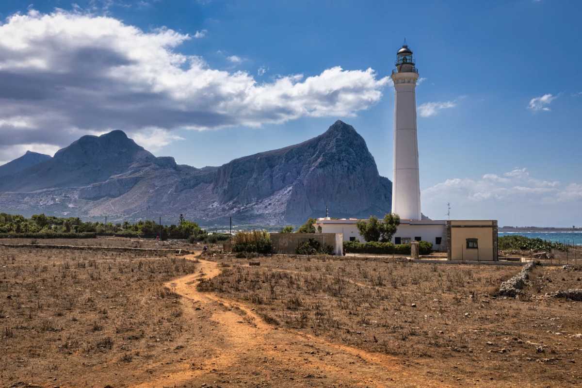 Primo piano della lanterna del Faro di San Vito Lo Capo al crepuscolo, con una coppia abbracciata in silhouette e le luci della città che iniziano ad accendersi
